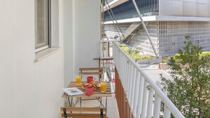 A balcony with a table set for breakfast, including orange juice and pastries, overlooking a modern building and a green area. - Acropolis Hideaway Studio by GHH (Athina)