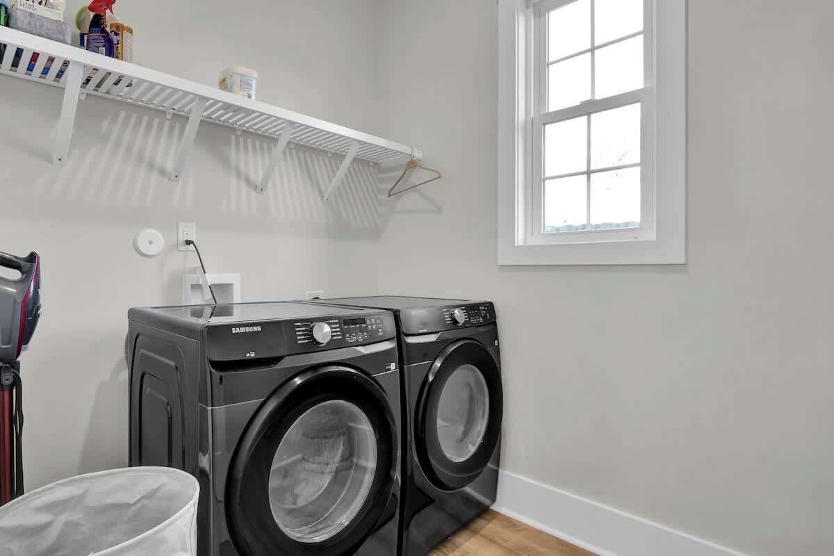 A laundry area with two Samsung washing machines, a white shelf with cleaning supplies, a white wall, and a window.