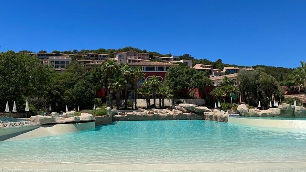 A resort pool area with clear blue water, surrounded by palm trees and rocks, and buildings in the background. - Air-conditioned apartment with sea view - Les Restanques SWIMMING POOLS TENNIS (Grimaud)