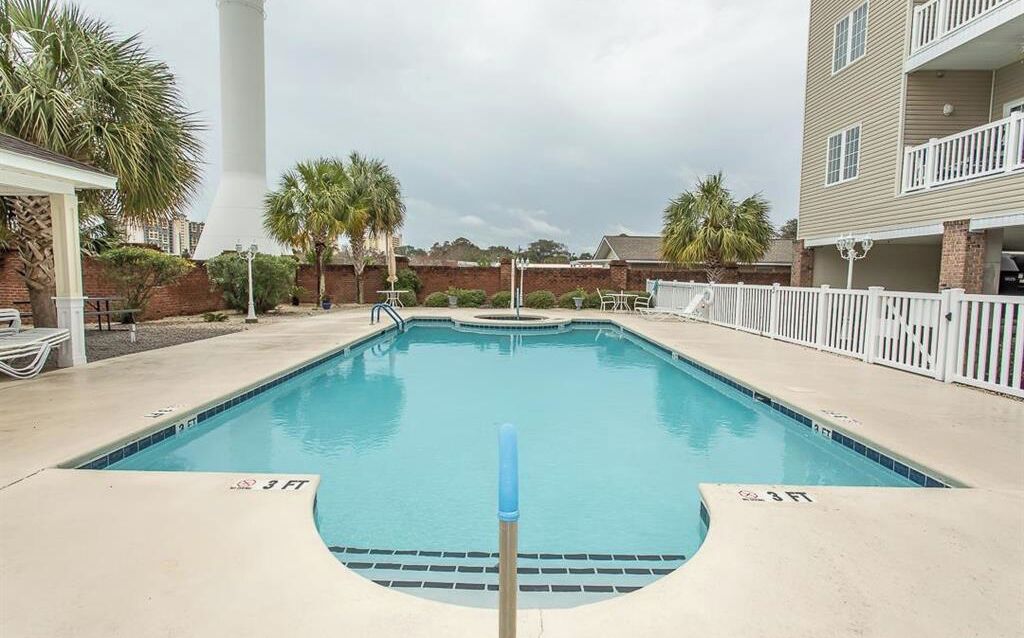 A swimming pool with a 3 FT depth marker, surrounded by a white fence, palm trees, and a brick wall.