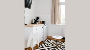 Una cocina compacta con muebles blancos, un televisor de pantalla plana negro montado en la pared, una pequeña mesa redonda y una alfombra estampada.