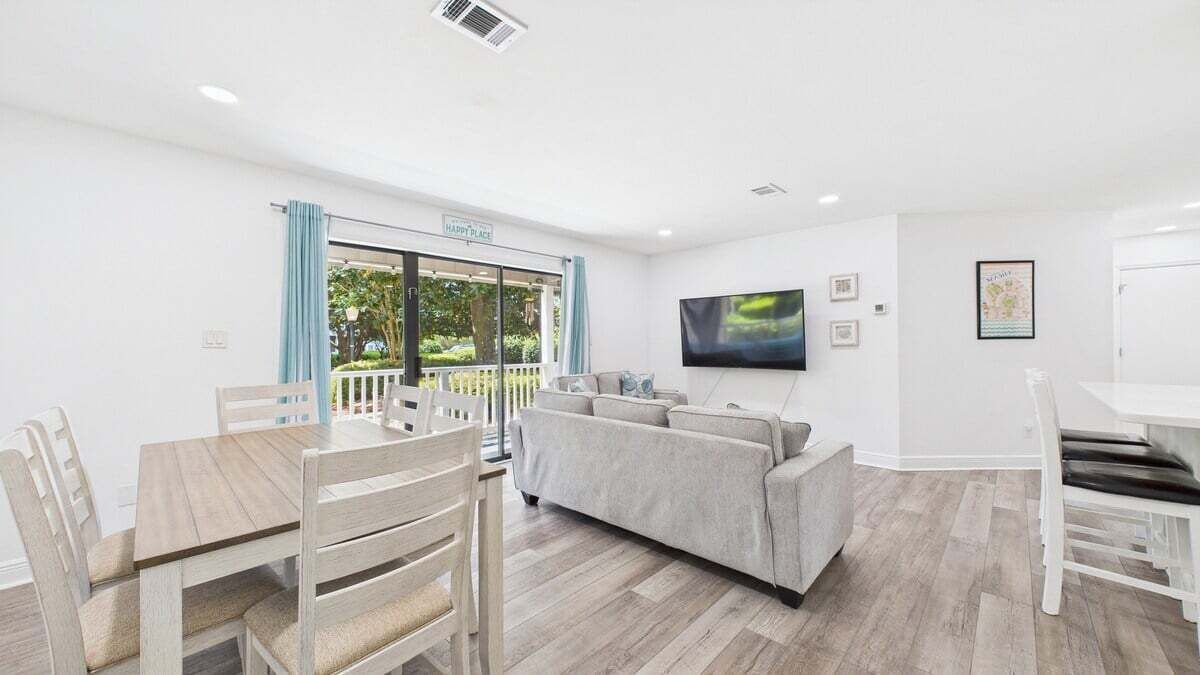 Un salon moderne avec un canapé gris, une table à manger en bois et des chaises. Un téléviseur à écran plat est fixé au mur.