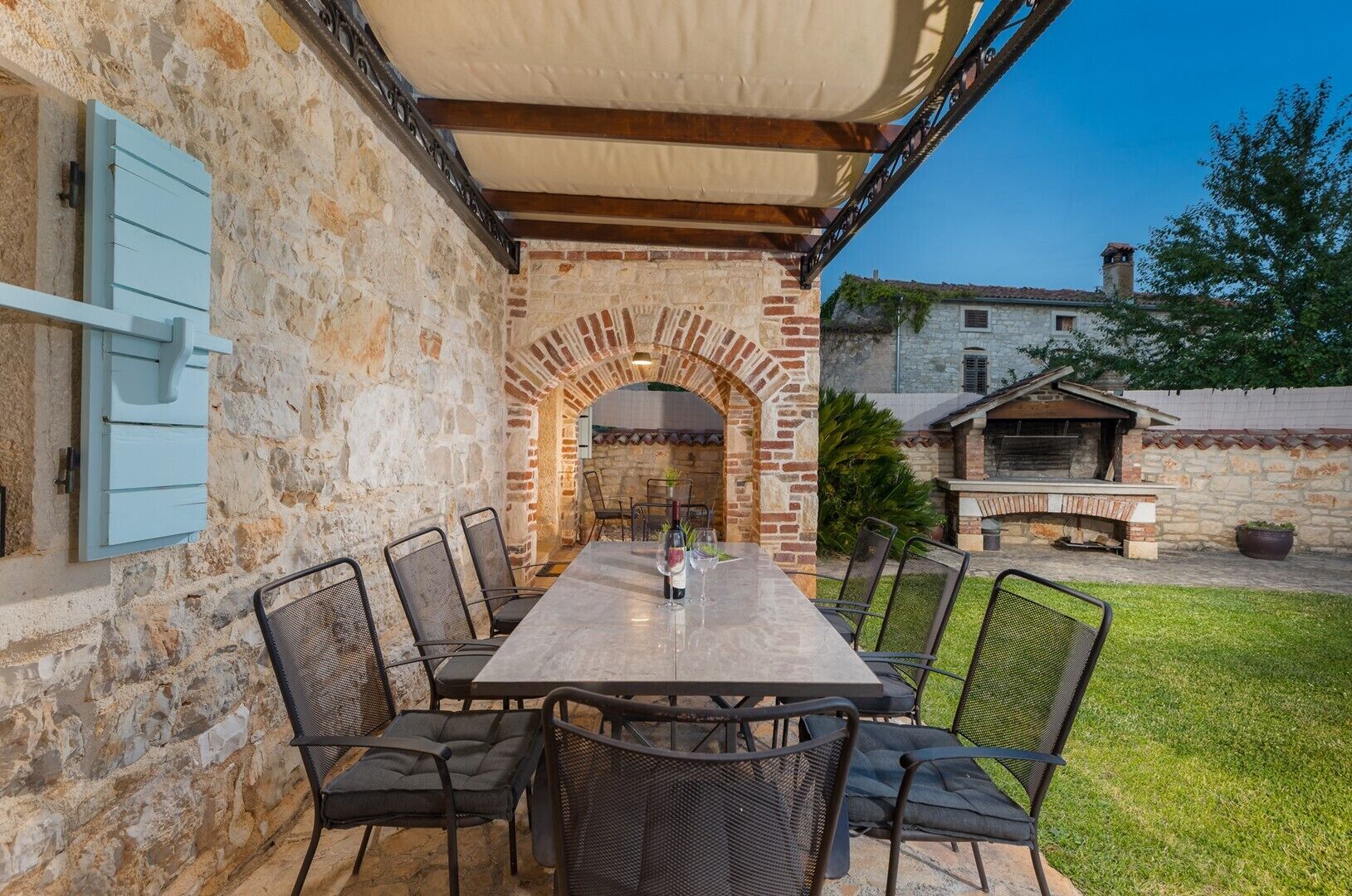 Un patio in pietra con un tavolo lungo e sedie, un arco e un forno a mattoni sullo sfondo.