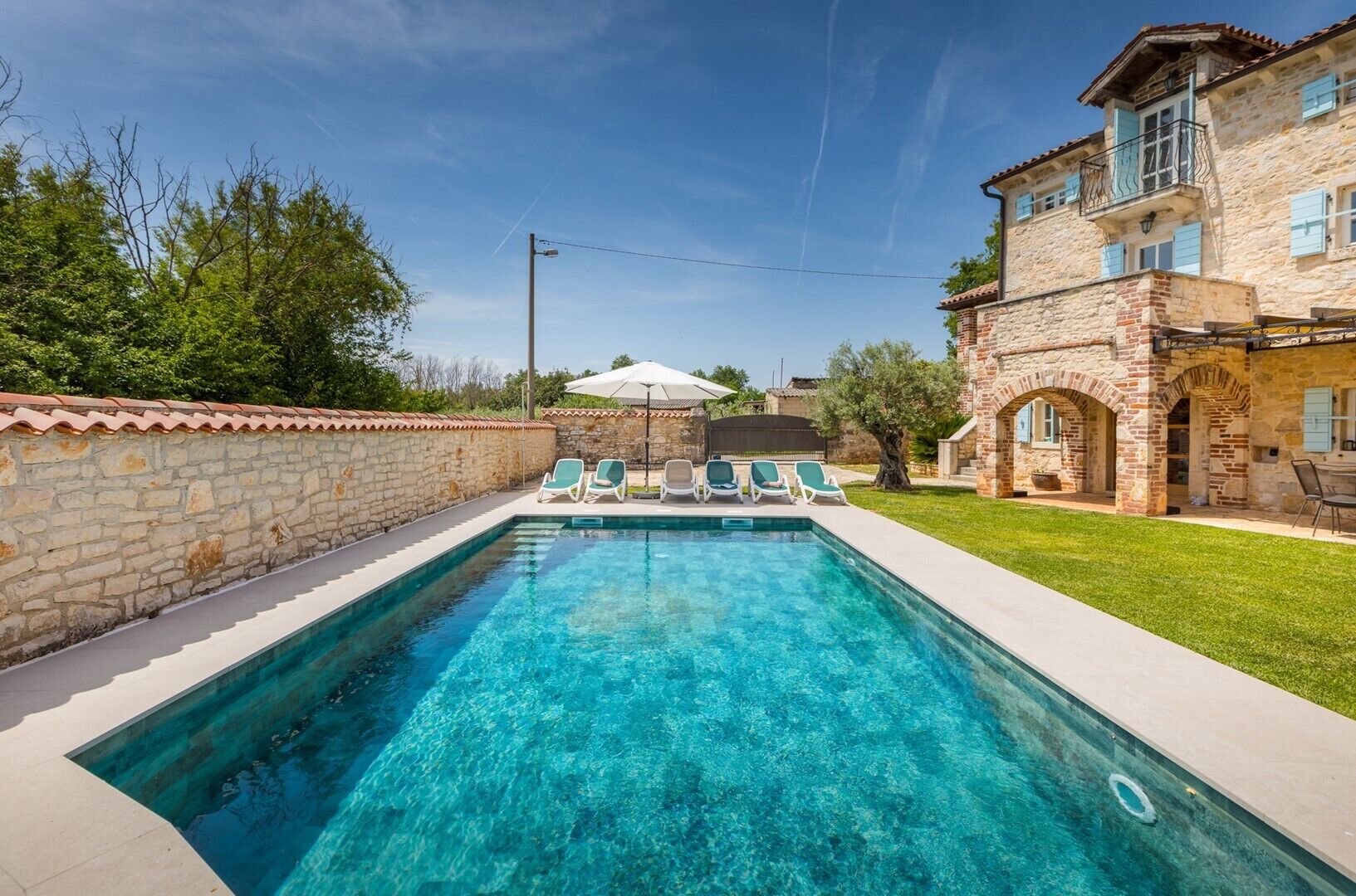 A rectangular swimming pool with clear blue water, surrounded by a stone wall and a grassy lawn. There are four lounge chairs aligned along the pool's edge.