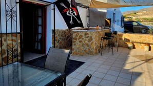 An outdoor patio with a stone wall, a glass table, and a flag. - Casita-1 To 5guests-pool-patio-bbq-parking (Huercal-Overa)