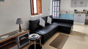 A living room with a black leather sofa, a small round table with a lamp, and a kitchen area with cabinets and a window. - Casita-1 To 5guests-pool-patio-bbq-parking (Huercal-Overa)