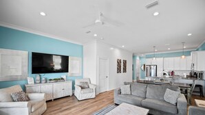 A modern living room with a large flat-screen TV, a grey sofa, a white armchair, and a dining area with a kitchen in the background. - Beach Facing, 4Bd, Sleeps 10, Bikes, BBQ, Pool (Gulf Shores)