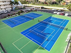 An aerial view of a tennis court complex with multiple courts, a parking area, and surrounding buildings.