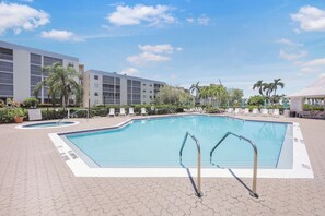 A swimming pool with a tiled deck, surrounded by lounge chairs and palm trees, with a building in the background. - Coastal Condo | Short Walk Away From the Beach (Marco Island)