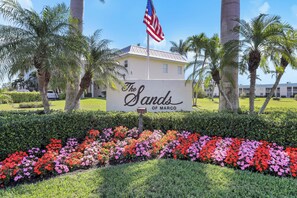 A sign for "The Sands of Marco" with a flag, surrounded by a flower bed and palm trees. - Coastal Condo | Short Walk Away From the Beach (Marco Island)