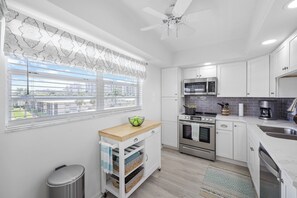 A modern kitchen with white cabinets, a microwave, an oven, and a sink. There's a window with blinds and a ceiling fan. - Coastal Condo | Short Walk Away From the Beach (Marco Island)