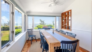 A dining area with a long wooden table, black chairs, and a ceiling fan. Large windows offer a view of the outdoors. - Paradise Awaits you!  Sleeping 14, this oceanfront complex is perfect! (Dennis Port)
