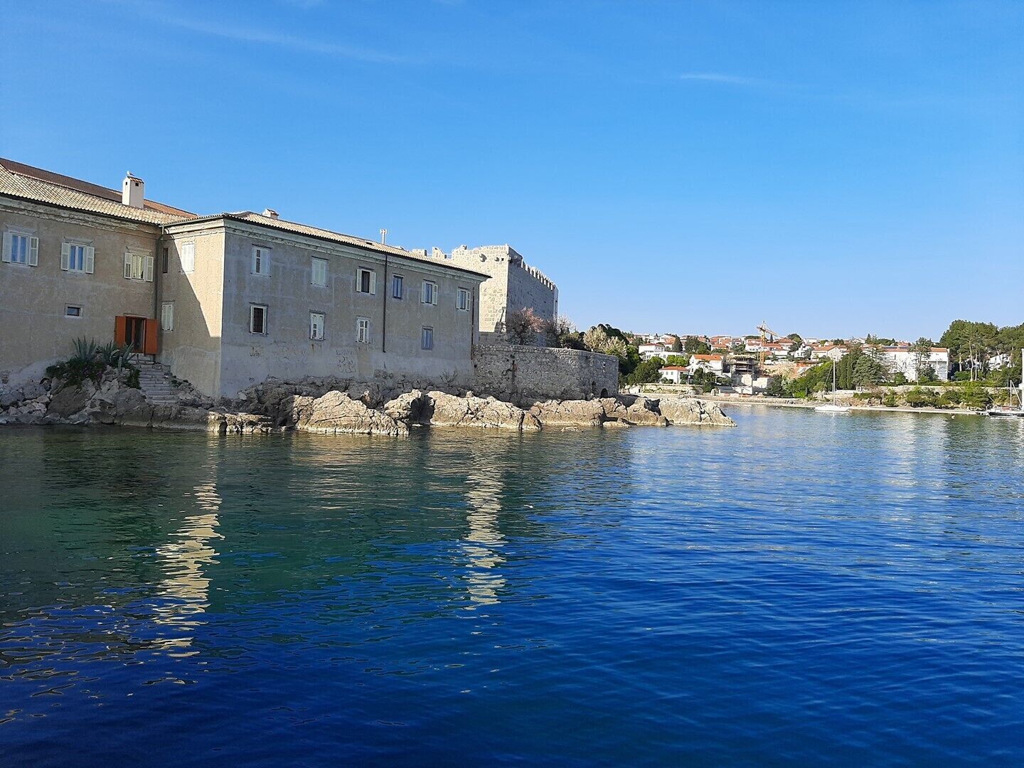 A historic stone building with a fortified wall by a calm body of water, likely a harbor or bay.