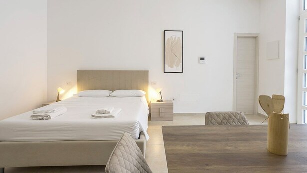 A modern bedroom with a large bed, bedside tables, a wooden floor, and a view of the outside. - Malcangi 29 (Trani)