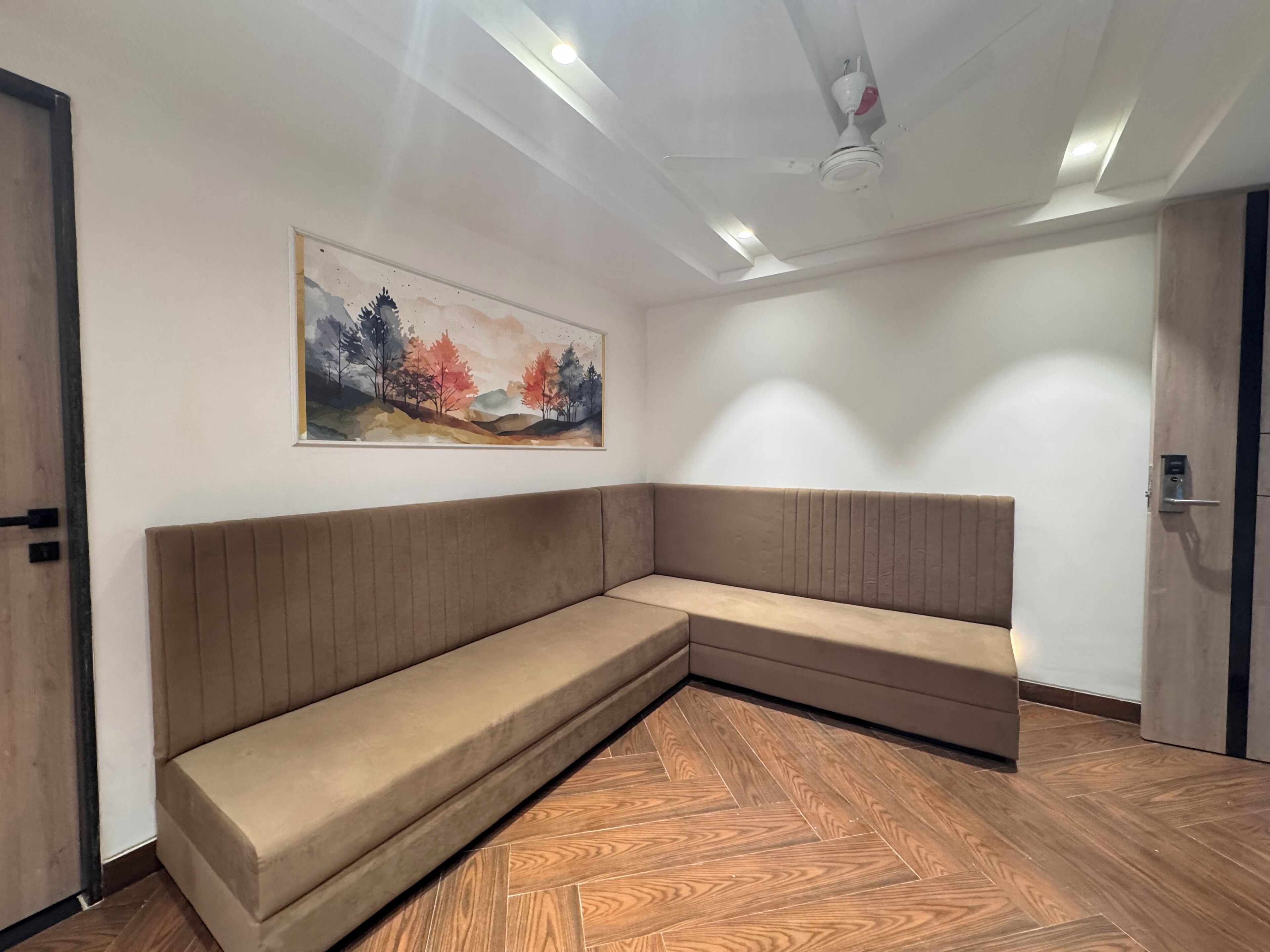 Una habitación moderna con un sofá seccional, suelo de madera en espiga, ventilador de techo, iluminación empotrada y un cuadro de paisaje en la pared.