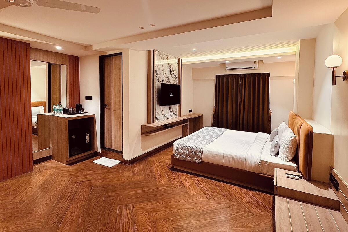 A hotel room with a bed, a desk, a mirror, a television, and an air conditioning unit.
