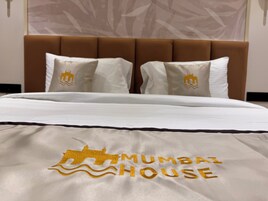 Una cama con una colcha blanca con un logo bordado en dorado y el texto "MUMBAI HOUSE".