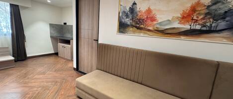 A modern living room with a sofa, a painting of autumn trees, and a kitchen area with a wooden cabinet and a countertop.