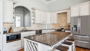 Una cocina con armarios blancos, una isla de granito con taburetes de bar, electrodomésticos de acero inoxidable y un salpicadero de azulejos.