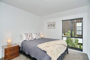 Un dormitorio con cama, mesita de noche, cómoda, ventana con vistas a la vegetación y una planta colgante en la pared.