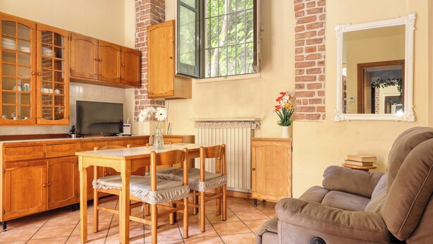 A cozy living room with a brown sofa, wooden dining table, and chairs. A TV is mounted on the wall, and there is a window with a view of trees. - JOIVY Charming flat in Porta Romana (Milan)