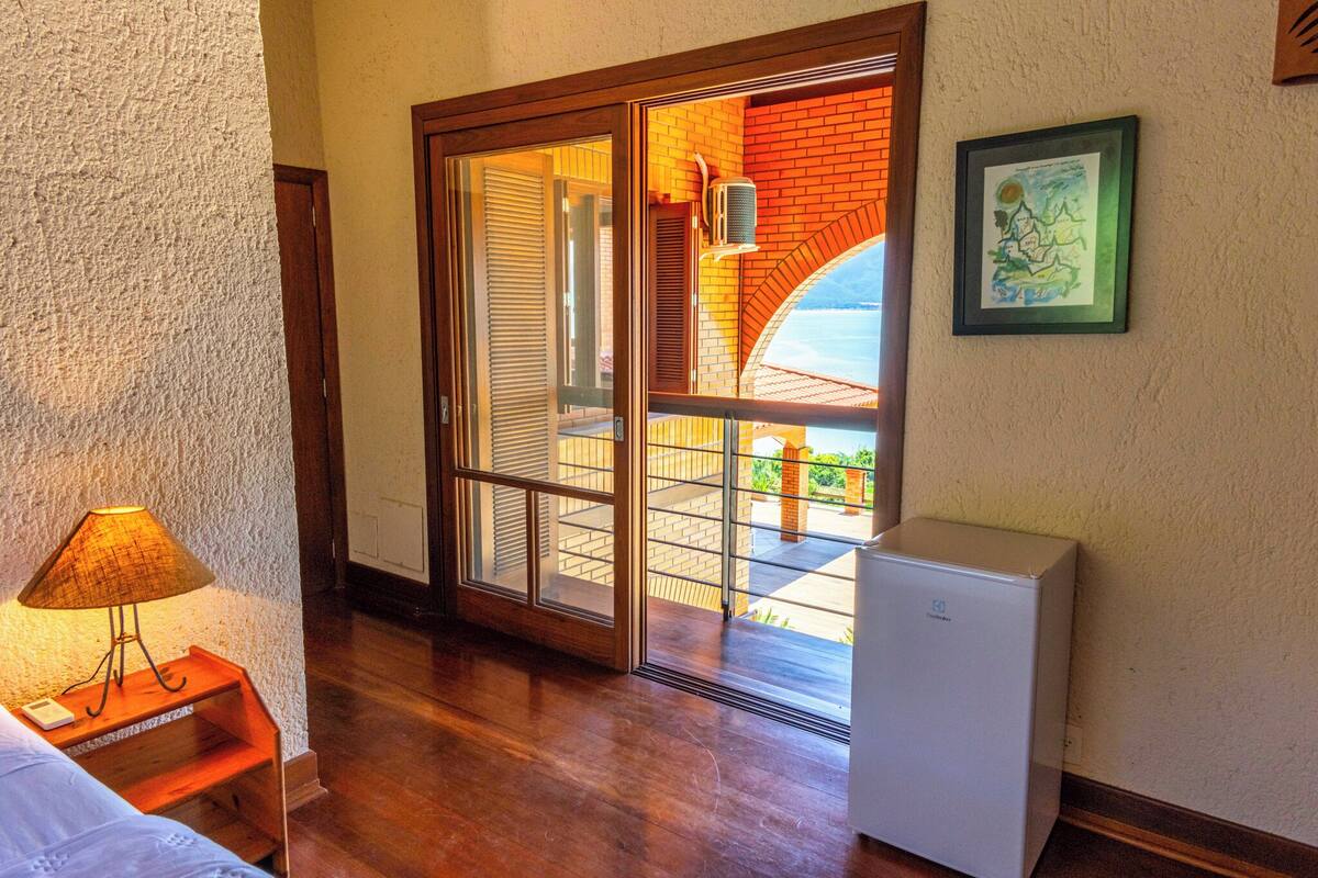 A room with a wooden floor, a bed, a nightstand with a lamp, a white refrigerator, and a sliding glass door leading to an outdoor area with a brick wall and a view of the sky.