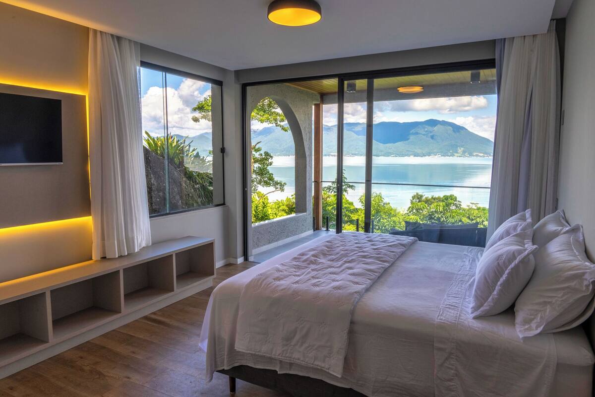 A bedroom with a large bed, a TV mounted on the wall, and a view of a lake and mountains through large windows.