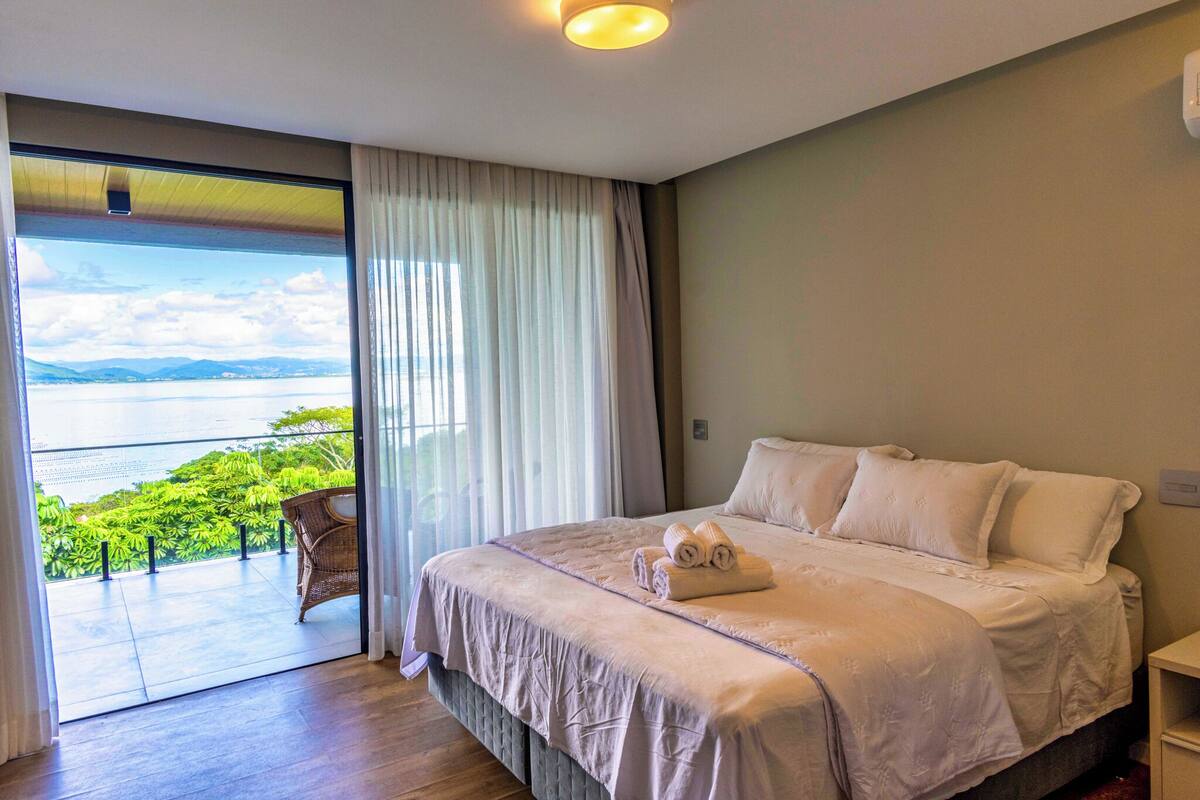 A hotel room with a large bed, a view of greenery and water through a sliding glass door, and a wicker chair by the door.