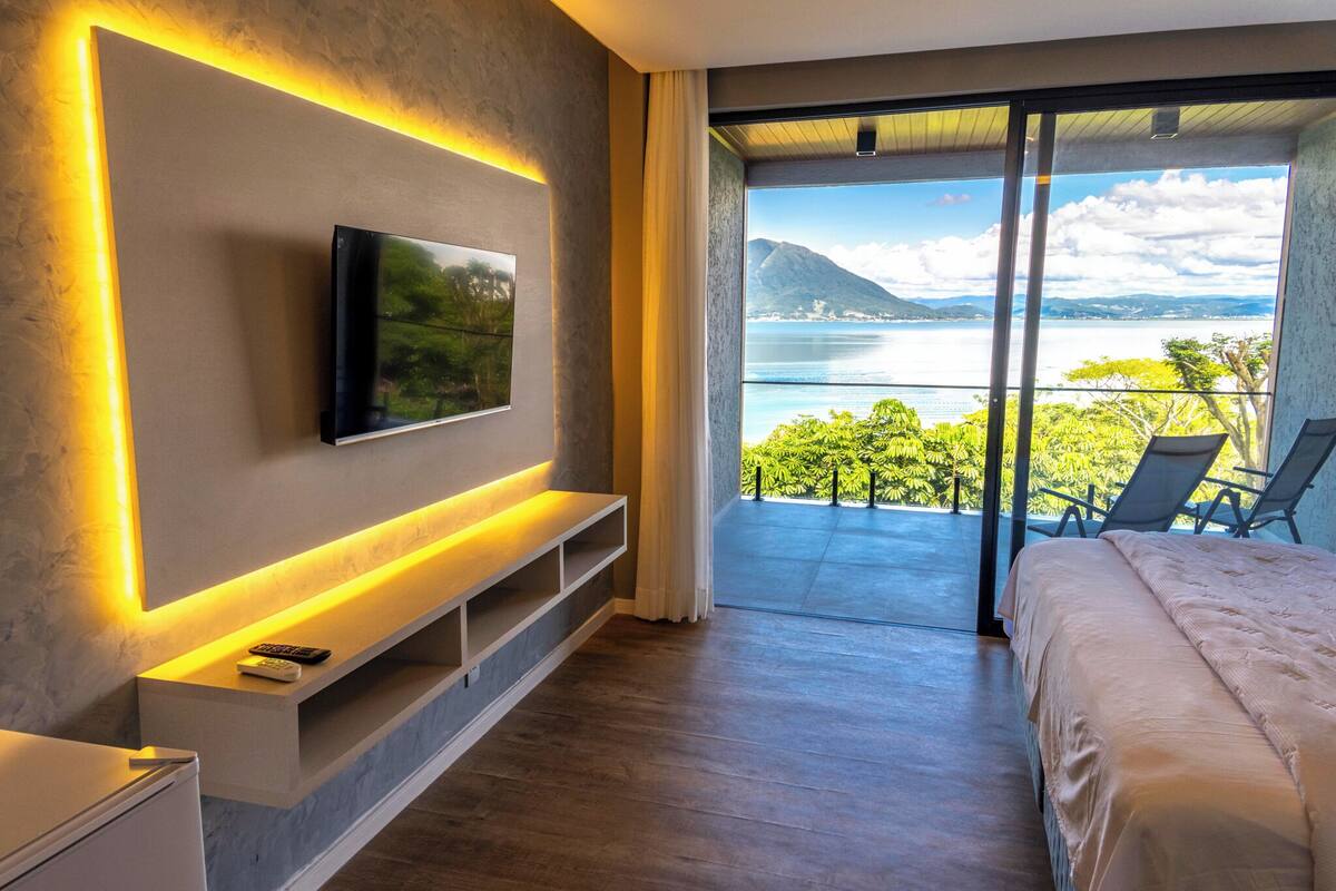 A modern bedroom with a TV mounted on the wall, a bed, a small desk, and a balcony with a view of the sea and mountains.