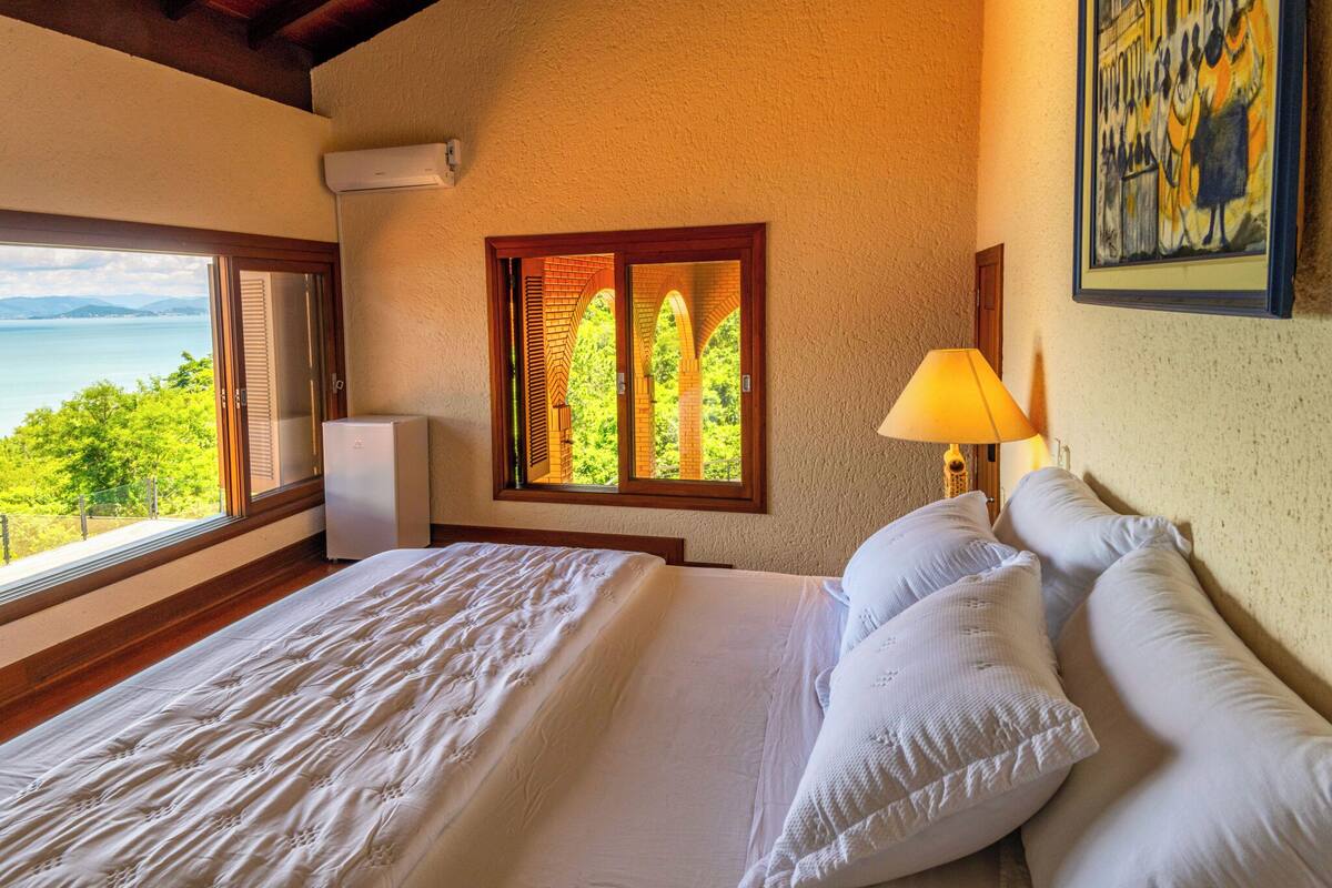 A bedroom with a large bed, a window with a view of greenery and a body of water, and a painting on the wall.