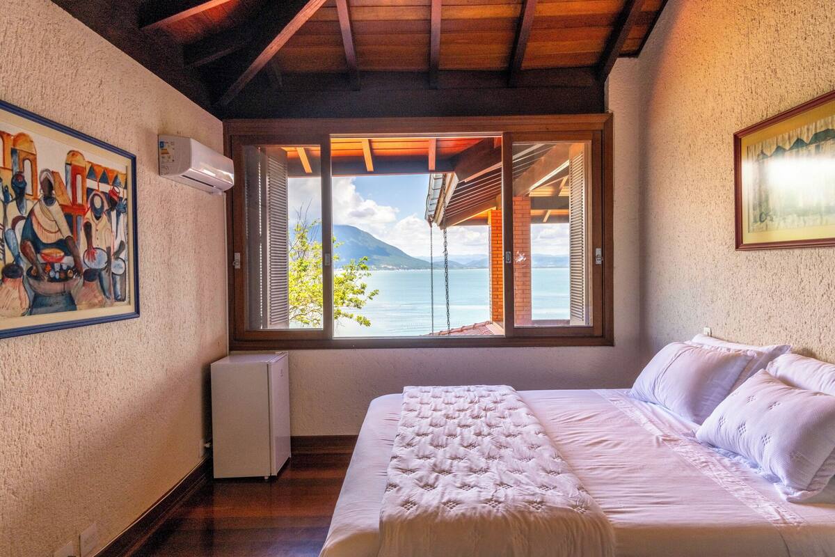 A bedroom with a large window offering a view of a lake and mountains.