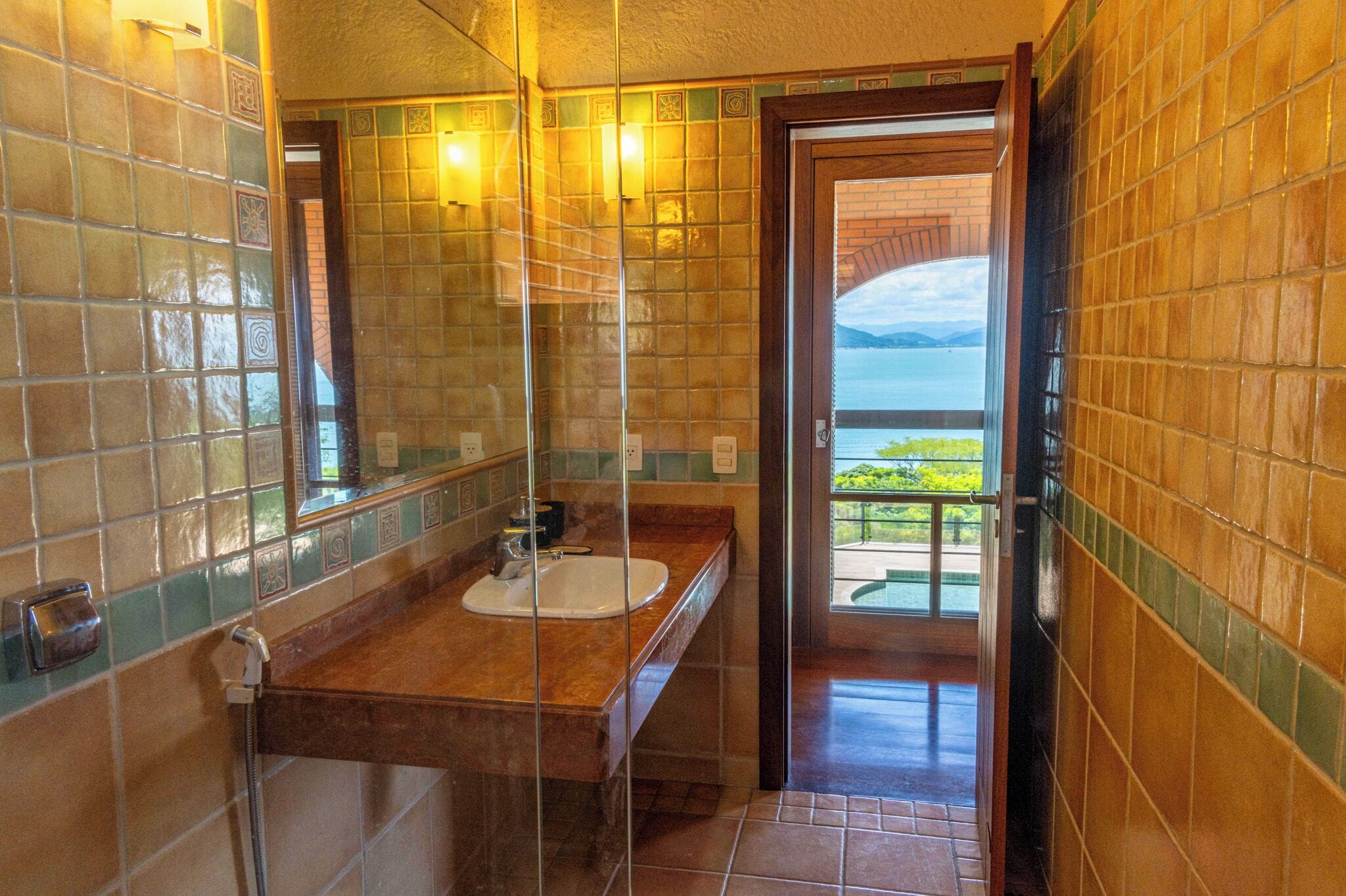 Een badkamer met een grote spiegel, een wastafel en een douche. De muren zijn betegeld met een mozaïekpatroon.