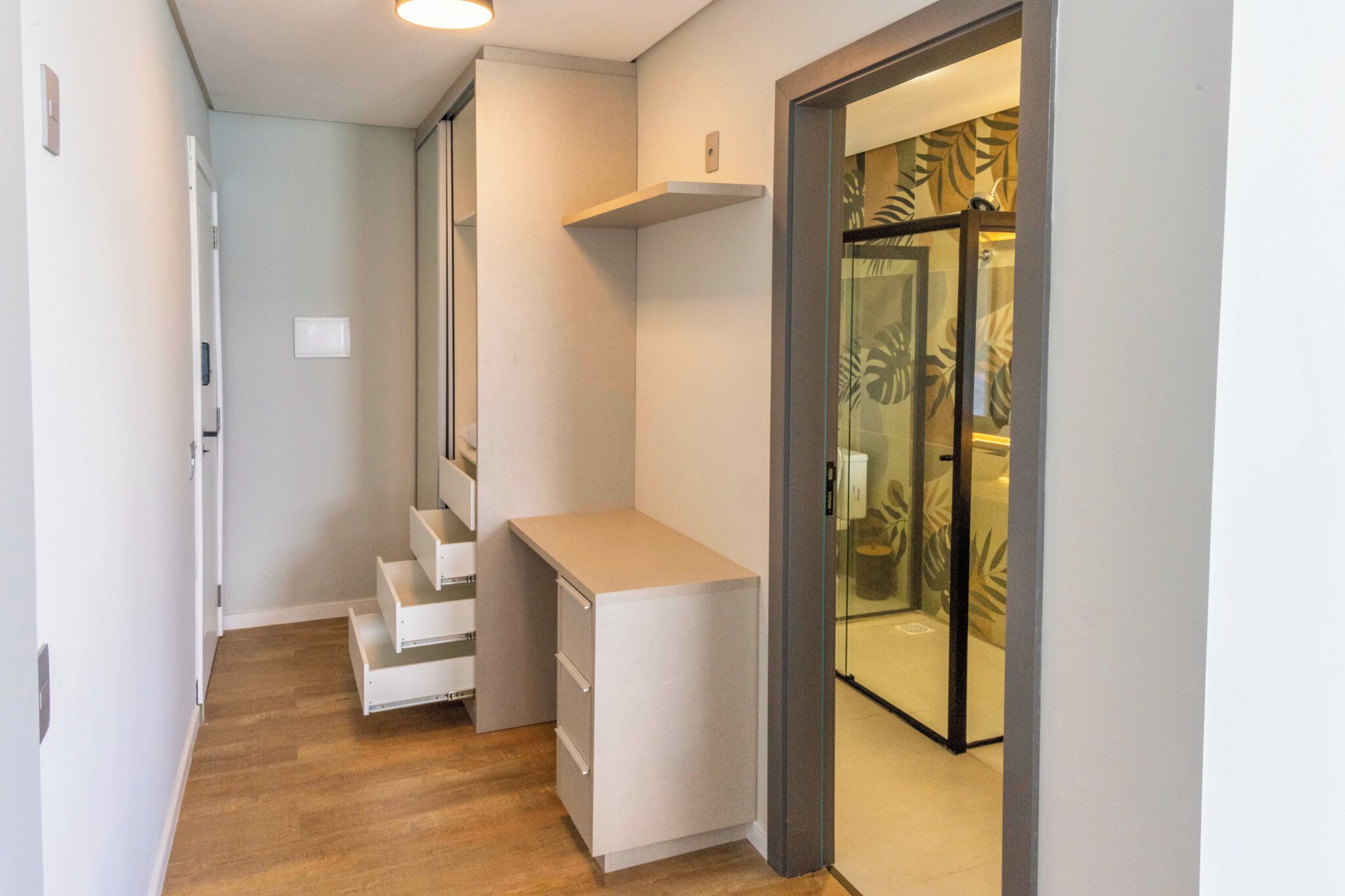 A narrow hallway with a white desk, a small seating area, and a glass door leading to a room with a tropical leaf pattern.