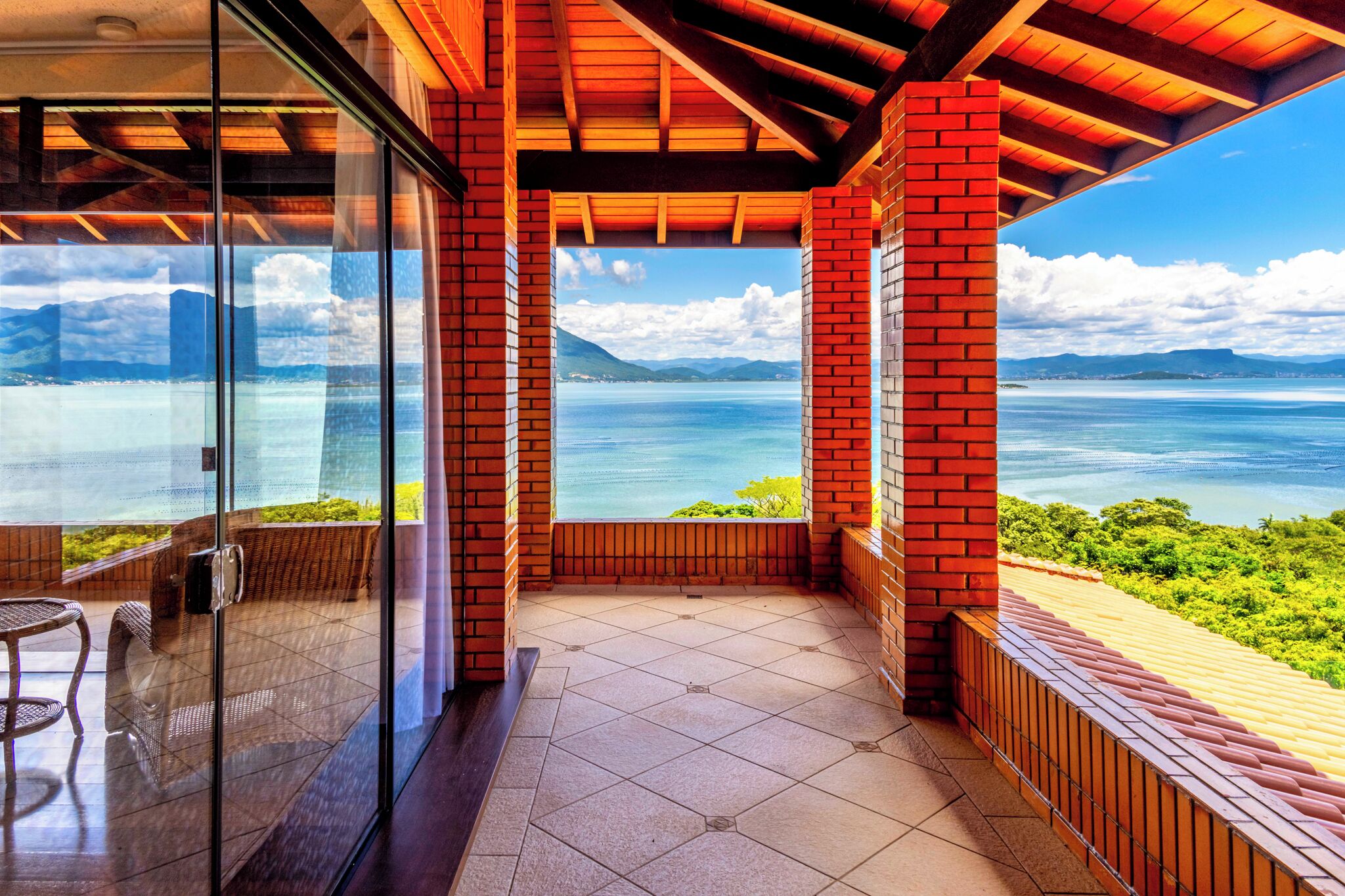 Uma varanda com vista para o mar e montanhas, com porta de vidro, mobiliário de madeira e chão em mosaico.