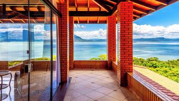 Uma varanda com vista para o mar e montanhas, com porta de vidro, mobiliário de madeira e chão em mosaico.