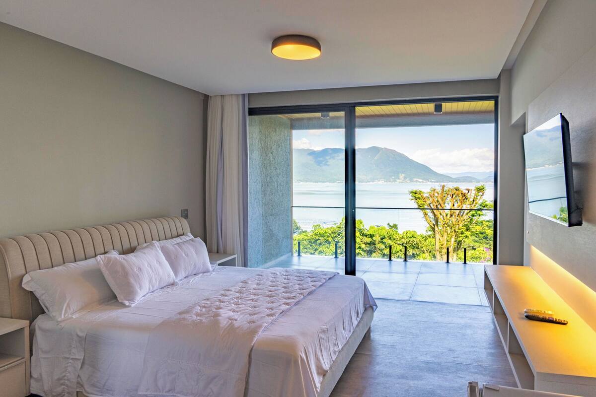 A modern hotel room with a large bed, a desk with a remote control, and a balcony with a view of the sea and mountains.