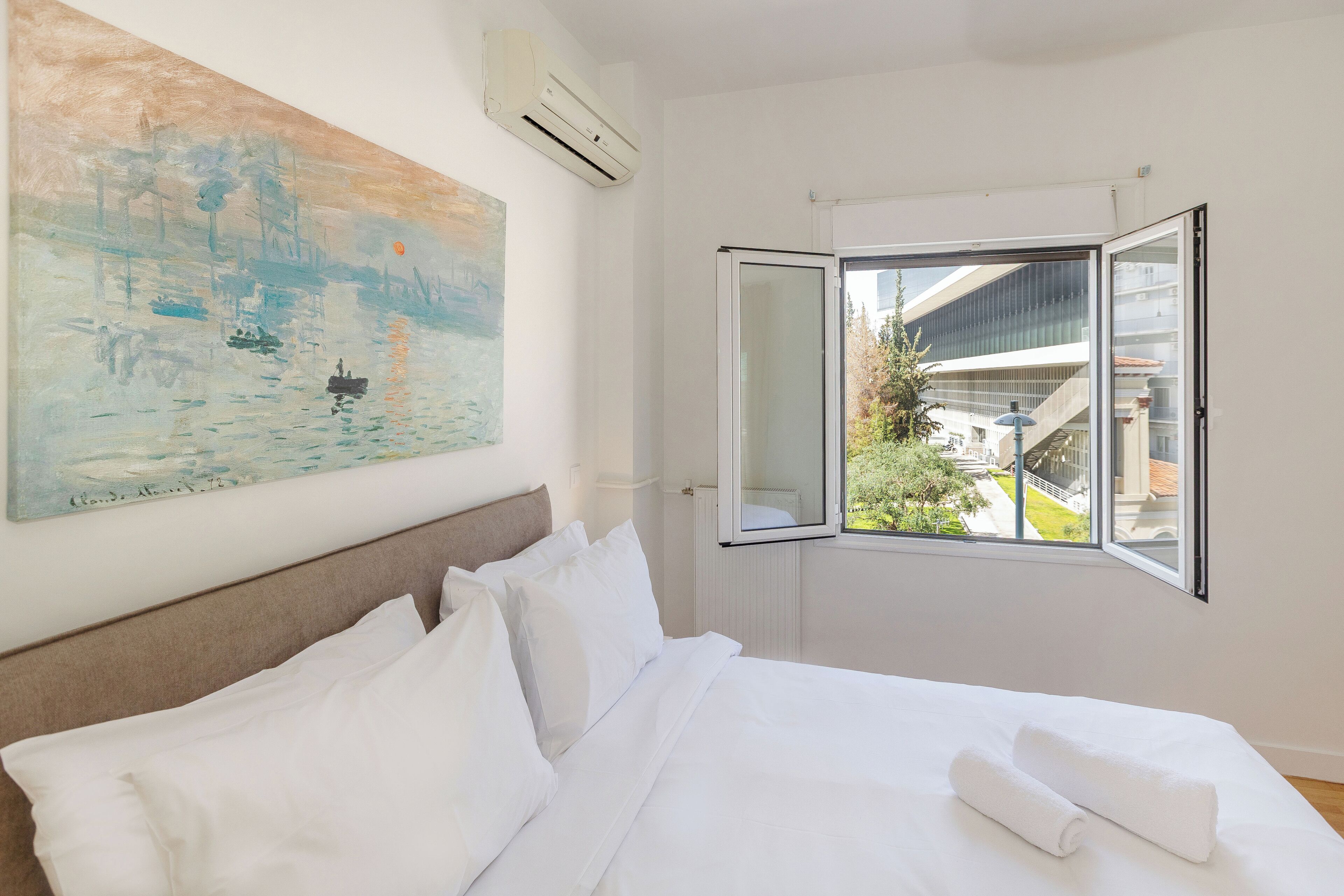 Quarto de hotel com cama, roupa de cama branca, um quadro na parede e janela com vista para edifícios e vegetação.