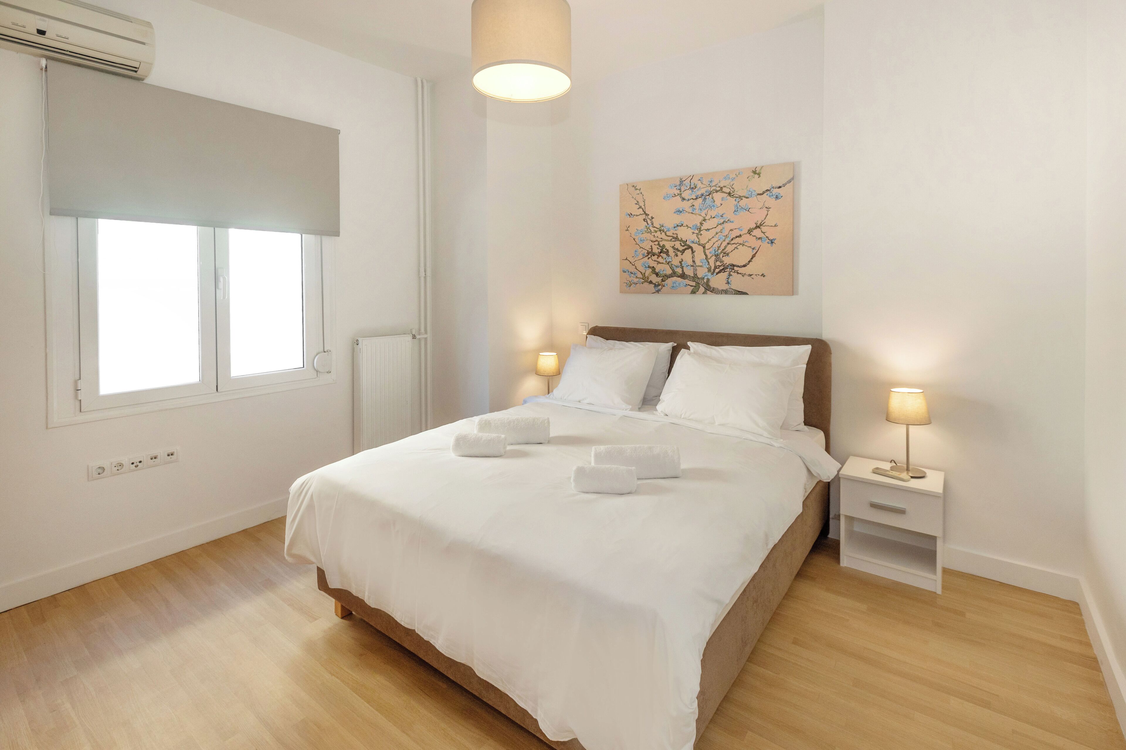 A bedroom with a bed, bedside tables, a lamp, a painting, and a window with blinds.