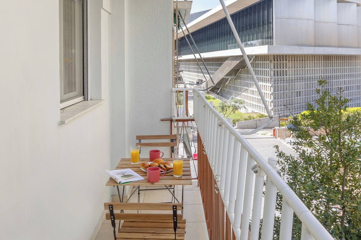 A balcony with a table set for breakfast, including orange juice and pastries, overlooking a modern building and a green area.