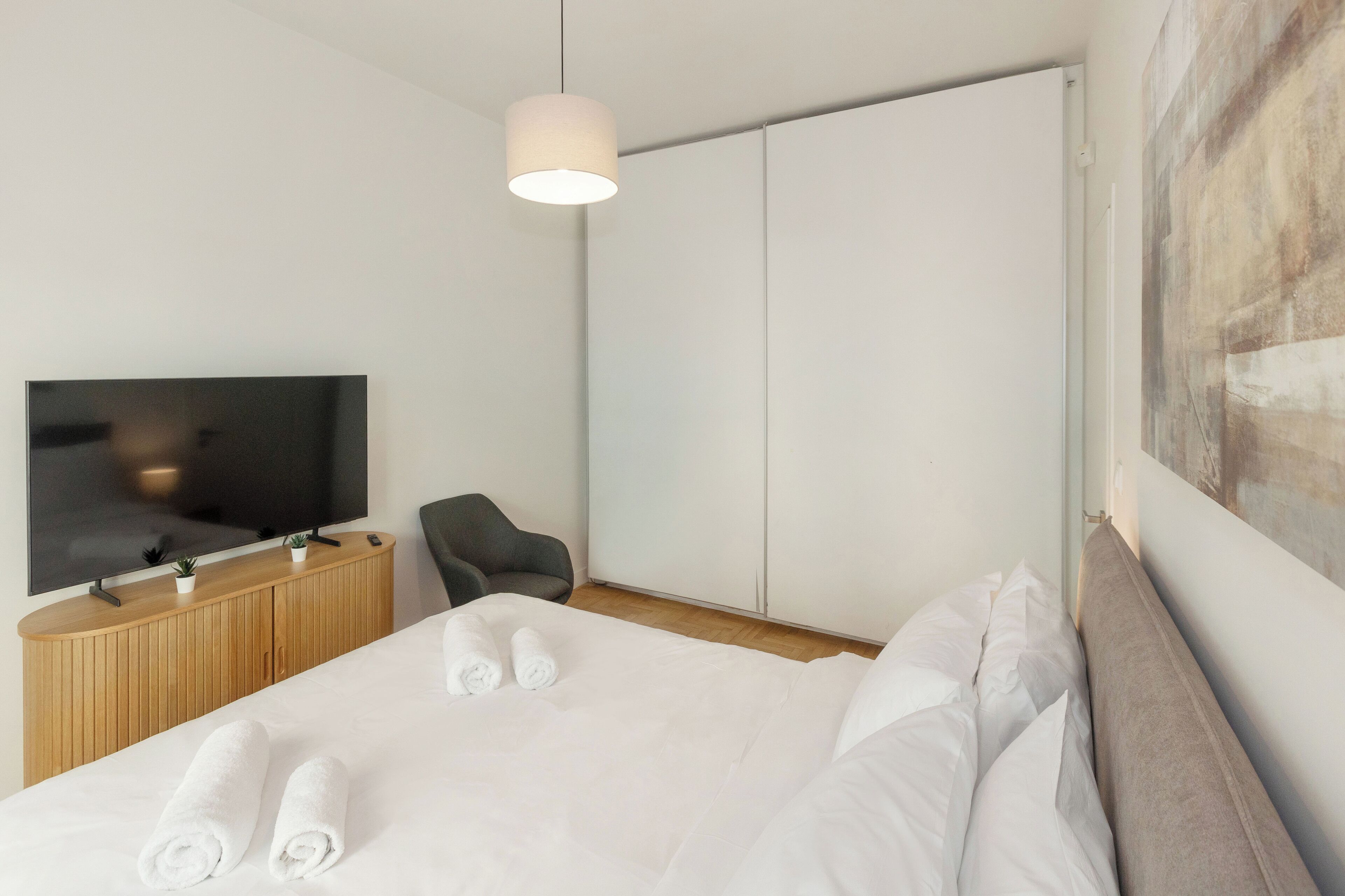 Une chambre d’hôtel moderne avec un grand lit, un téléviseur à écran plat, une console en bois, une chaise et un tableau au mur.