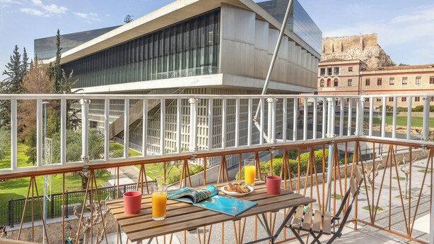 A balcony with a table set for breakfast, overlooking a modern building and a historic structure. - Acropolis Museum Retreat by GHH (Athens)