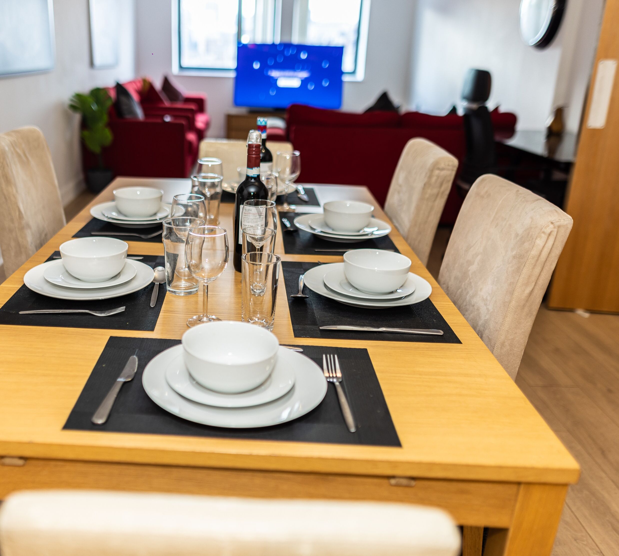 Ein Esszimmer mit Esstisch, gedeckten weißen Schüsseln, Gläsern und Besteck, einer Weinflasche und einem Fernseher im Hintergrund.