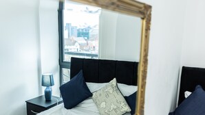 A bedroom with a bed, a nightstand, a lamp, and a window with a view of buildings. - Apartment Sleeps 6 With Balcony and Parking (Birmingham)