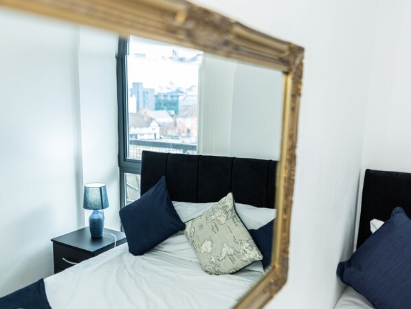 A bedroom with a bed, a nightstand, a lamp, and a window with a view of buildings. - Apartment Sleeps 6 With Balcony and Parking (Birmingham)