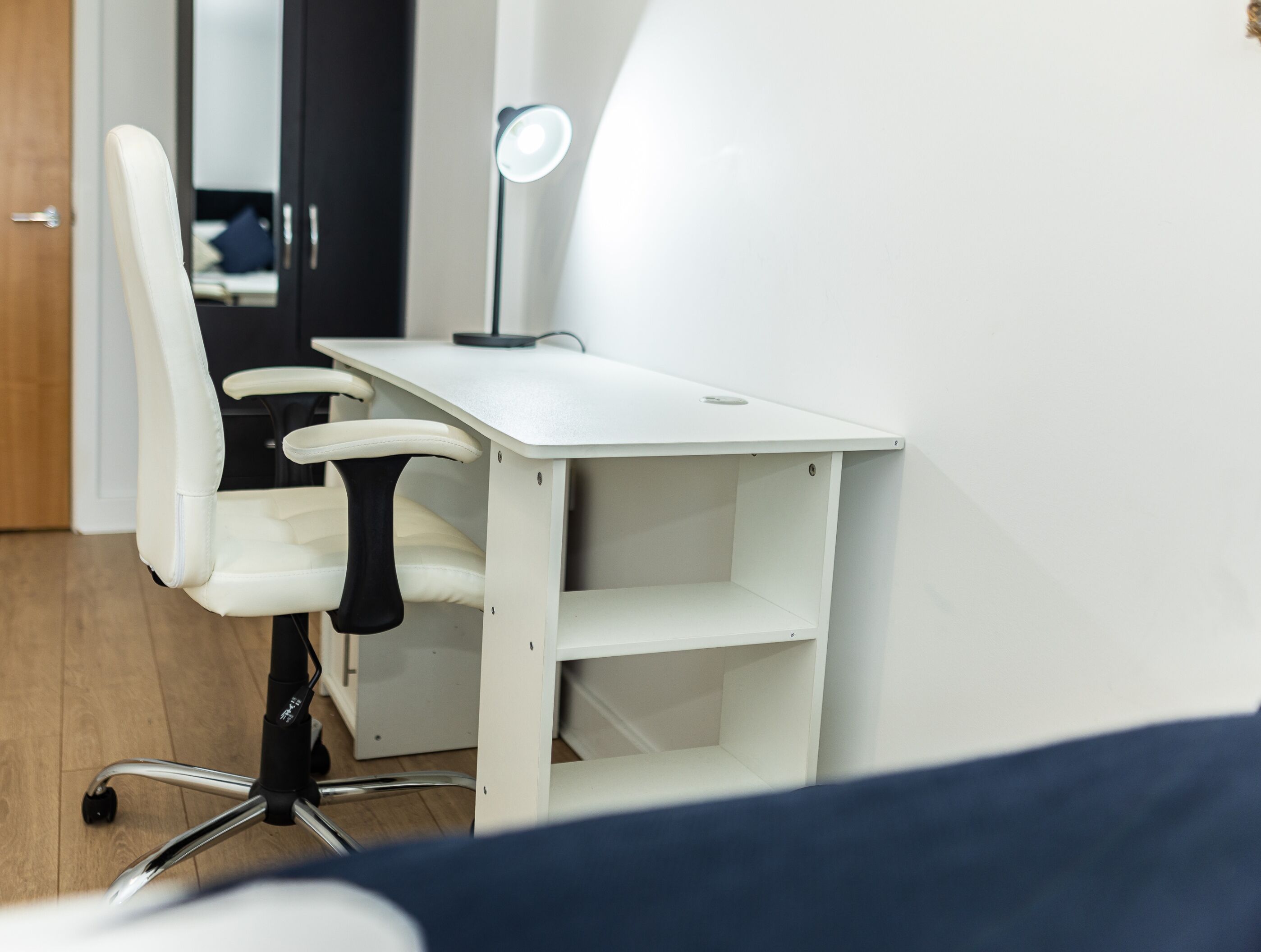 Een wit bureau met een lamp, een stoel en een spiegel in een kamer met houten vloer en een deur.