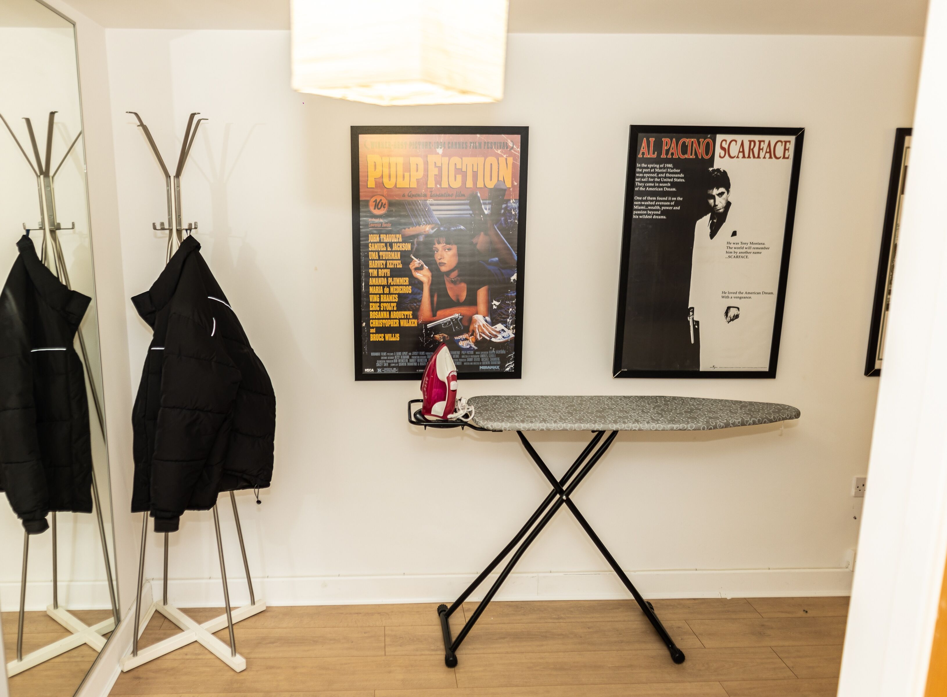 A room with a standing mirror, a coat rack with two black coats, an ironing board, and two posters on the wall.