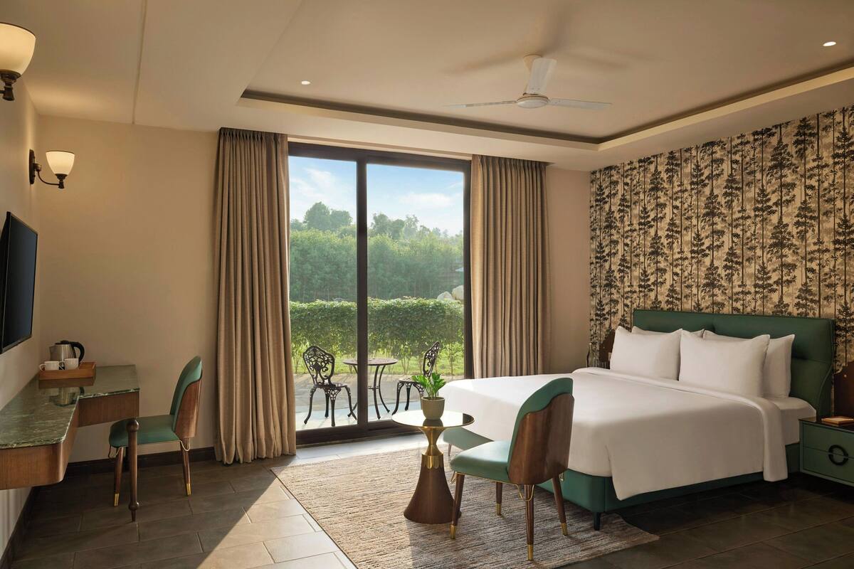 A hotel room with a bed, desk, chairs, and a view of greenery.