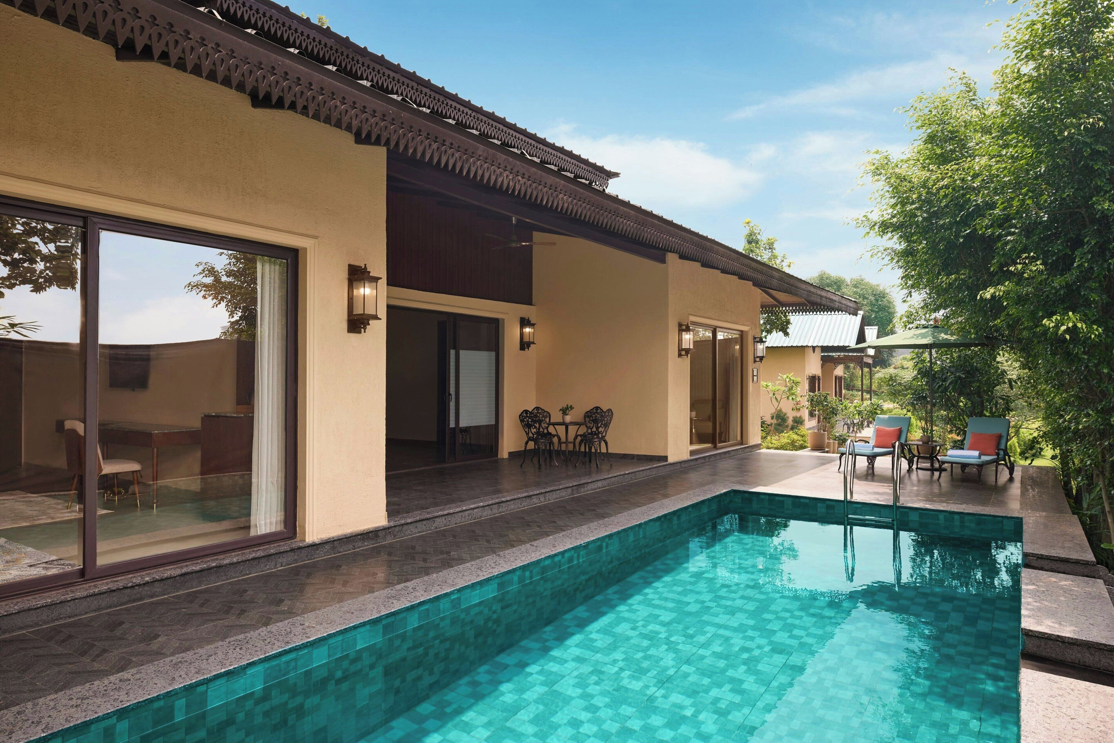 A swimming pool with a tiled edge, a covered patio area with outdoor seating, and a building with large sliding glass doors.