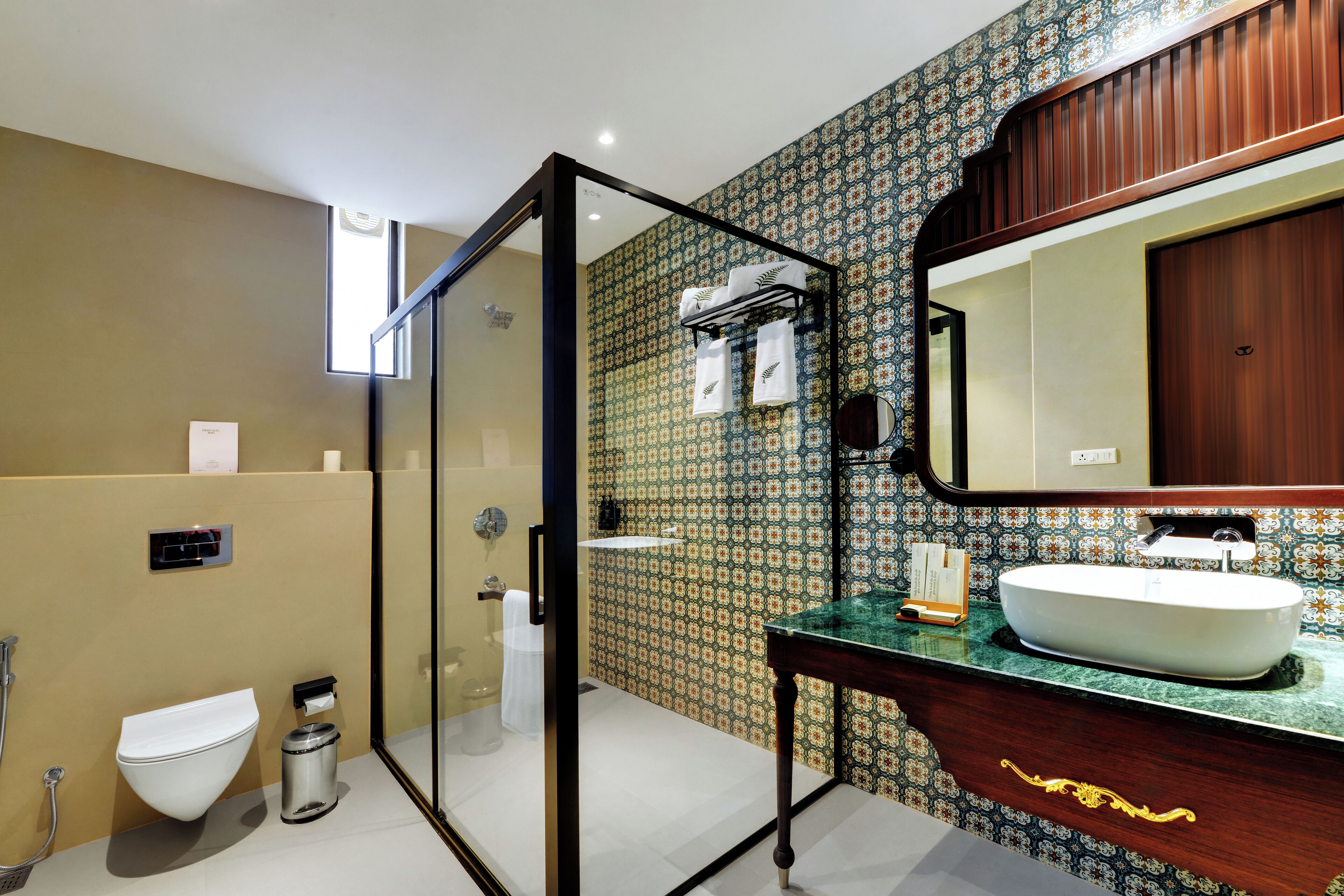 Un baño con cabina de ducha de vidrio, inodoro blanco, lavamanos con encimera verde y papel tapiz estampado.