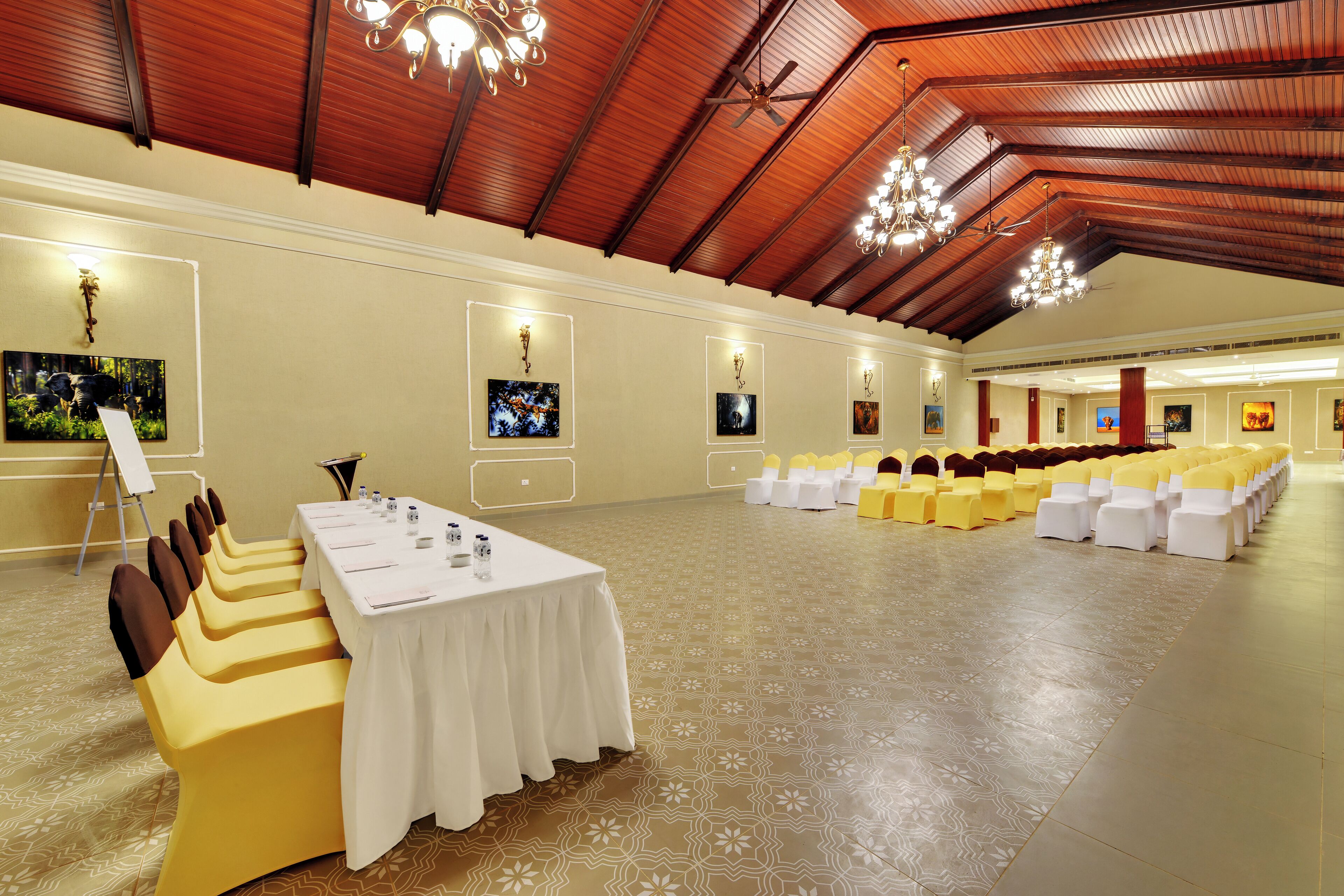A spacious hall with a high ceiling, rows of yellow and white chairs, a long table with a white tablecloth, and framed pictures on the walls.
